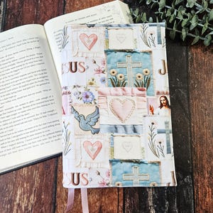 Op de afbeelding: Een stoffen boekomslag met een patchwork ontwerp met harten, kruisen, bloemen en een duif. De omslag heeft het woord "JESUS" en een afbeelding van Jezus. Een open boek ligt ernaast.