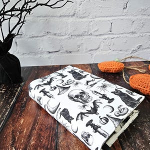 May include: A white book cover with a black and white Halloween themed print. The print features bats, skulls, pumpkins, cats, snakes, and other spooky imagery.