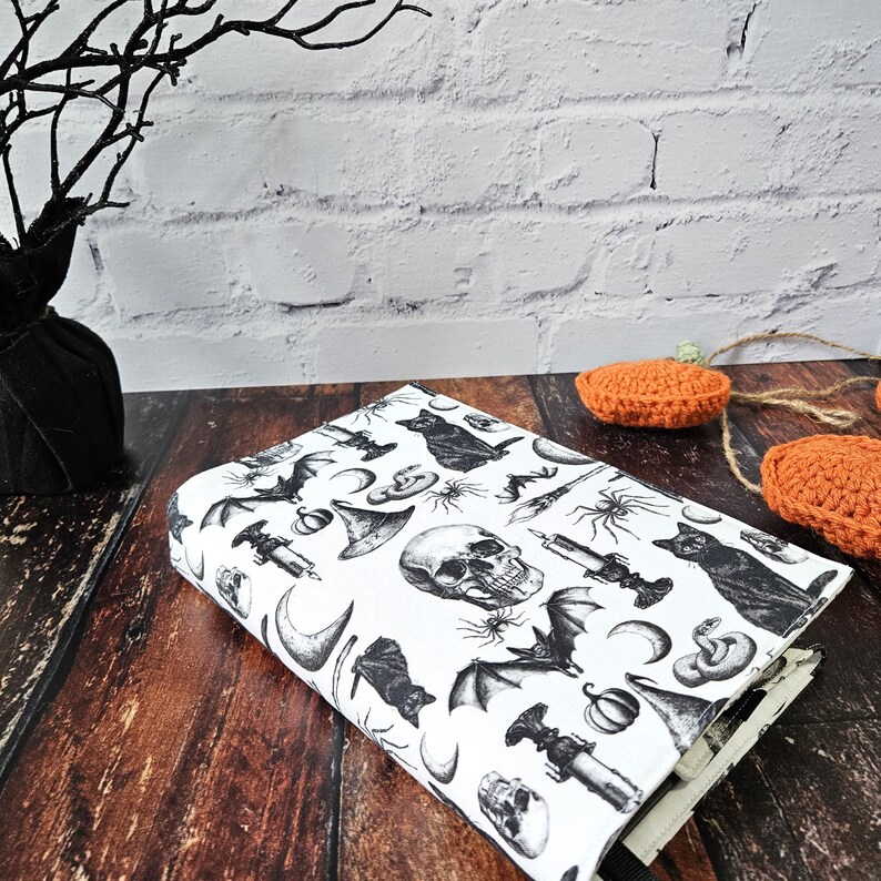 May include: A white book cover with a black and white Halloween themed print. The print features bats, skulls, pumpkins, cats, snakes, and other spooky imagery.