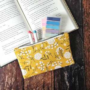 May include: A yellow zippered pouch with a white floral pattern. The pouch is lying on a wooden surface with a book and sticky notes.