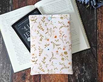 May include: A pink and white floral patterned e-reader sleeve with a blue button closure. The sleeve is lying on a wooden surface next to a book titled "A Court of Thorns and Roses".