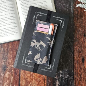 Puede incluir: Un cuaderno negro con un bolsillo de tela negro y gris sujeto a la portada delantera. El bolsillo contiene un bolígrafo blanco y notas adhesivas rosas. El cuaderno tiene un borde decorativo alrededor de los bordes.