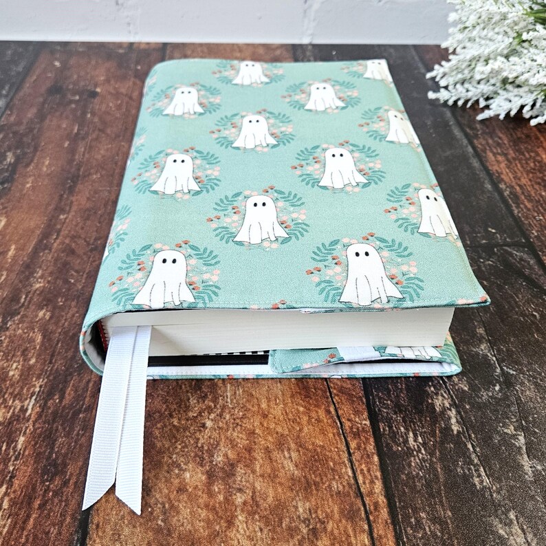 Peut inclure: Une housse de livre turquoise avec un motif de fant&ocirc;mes blancs. Les fant&ocirc;mes sont entour&eacute;s de petites couronnes de feuilles et de fleurs. La housse est munie d'un marque-page blanc.