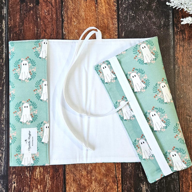 Peut inclure: Une couverture de livre en tissu vert avec un motif de fant&ocirc;mes blancs et une fermeture en ruban blanc. La couverture est ouverte pour r&eacute;v&eacute;ler un int&eacute;rieur en tissu blanc.