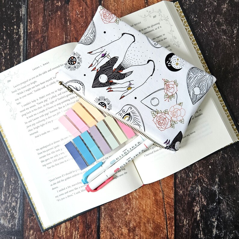 K&ouml;nnte beinhalten: Eine wei&szlig;e Rei&szlig;verschlusstasche mit einem schwarz-wei&szlig;en Druck von H&auml;nden, Augen, Rosen und einem Ouija-Brett. Die Tasche liegt offen auf einem Buch mit bunten Haftnotizen und rosa und blauen Markern.