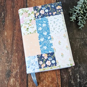 Pode incluir: Uma capa de livro patchwork com padrões florais em tons de rosa, azul e branco. Inclui uma fita de marcador azul claro. O design combina elementos florais e geométricos.