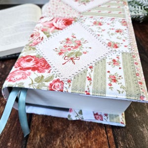K&ouml;nnte beinhalten: Ein Buchumschlag aus floralem Stoff mit Patchwork-Design. Der Umschlag zeigt eine Mischung aus Mustern, darunter Rosen und Streifen, in Rosa, Gr&uuml;n und Wei&szlig;. Ein hellblaues Lesezeichenband ist sichtbar. Das Buch ist ge&ouml;ffnet.