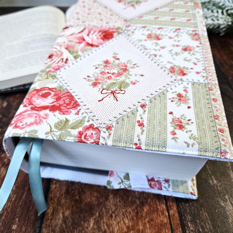 K&ouml;nnte beinhalten: Ein Buchumschlag aus floralem Stoff mit Patchwork-Design. Der Umschlag zeigt eine Mischung aus Mustern, darunter Rosen und Streifen, in Rosa, Gr&uuml;n und Wei&szlig;. Ein hellblaues Lesezeichenband ist sichtbar. Das Buch ist ge&ouml;ffnet.