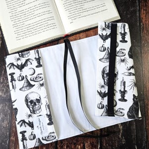 May include: A white book cover with black and white illustrations of bats, skulls, snakes, candles, and pumpkins. The cover has two black ribbon ties. The book cover is made by Elizabeth Maguire.
