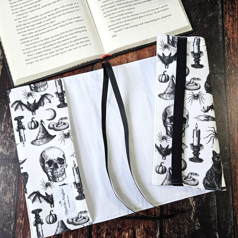 May include: A white book cover with black and white illustrations of bats, skulls, snakes, candles, and pumpkins. The cover has two black ribbon ties. The book cover is made by Elizabeth Maguire.