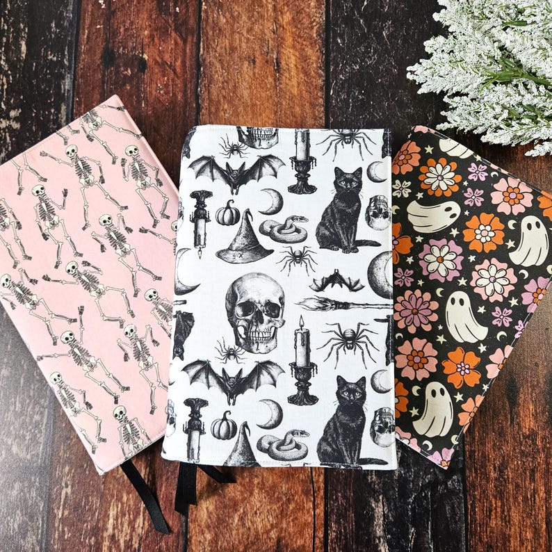 May include: Three Halloween-themed book covers. The first cover is pink with a white skeleton pattern. The second cover is white with a black skull, bat, cat, spider, snake, and candle pattern. The third cover is black with a white ghost and orange and pink flower pattern.