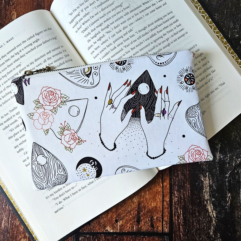 K&ouml;nnte beinhalten: Eine wei&szlig;e Rei&szlig;verschlusstasche mit einem sich wiederholenden Schwarz-Wei&szlig;-Muster von H&auml;nden, die ein schwarzes und wei&szlig;es Ouija-Brett mit einem Halbmond halten. Das Muster enth&auml;lt auch Rosen, Sterne und andere okkulte Symbole. Der Text "It's my burden to be... You don't have to... I do. What I have to face, what I survive." ist auf der Unterseite der Tasche aufgedruckt.