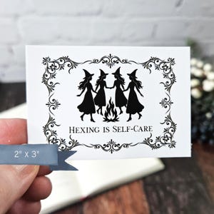 May include: A white card with a black and white illustration of three witches dancing around a fire. The text "Hexing is Self-Care" is printed below the illustration. The card is 2 inches by 3 inches.