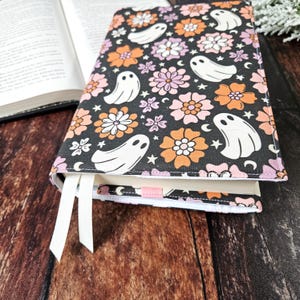 May include: A black book cover with a white ribbon bookmark. The cover features a repeating pattern of orange and pink flowers, white ghosts, and stars on a black background.
