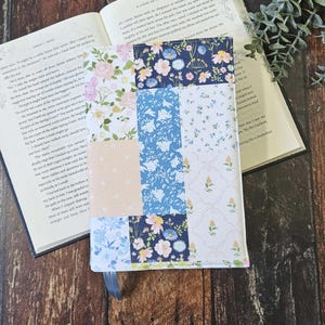 May include: A fabric book cover with a patchwork design featuring floral patterns in pink, blue, and white. It includes a blue ribbon bookmark. The open book displays text on its pages.