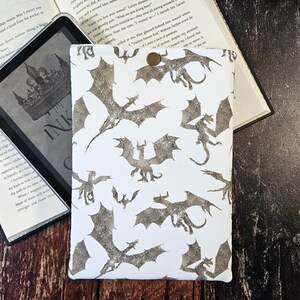 K&ouml;nnte beinhalten: Wei&szlig;er Kindle-Schutzbezug mit einem sich wiederholenden Muster aus silbernen Glitzerdrachen. Der Bezug hat einen Knopfverschluss.