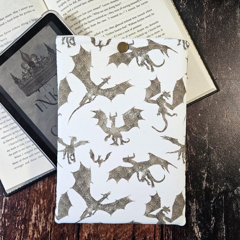 K&ouml;nnte beinhalten: Wei&szlig;er Kindle-Schutzbezug mit einem sich wiederholenden Muster aus silbernen Glitzerdrachen. Der Bezug hat einen Knopfverschluss.