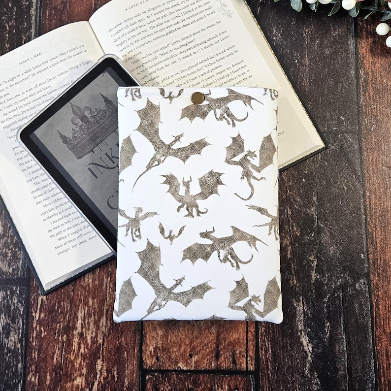 K&ouml;nnte beinhalten: Wei&szlig;e E-Reader-H&uuml;lle mit einem sich wiederholenden Muster aus silbernen Drachen. Die H&uuml;lle hat einen Knopfverschluss.