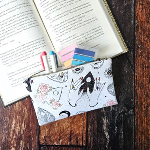 K&ouml;nnte beinhalten: Eine wei&szlig;e Rei&szlig;verschlusstasche mit einem schwarz-wei&szlig;en Druck von H&auml;nden, die ein schwarzes Dreieck mit einer Sichelmond und Sternen halten. Die Tasche ist mit einem sich wiederholenden Muster aus Augen, Rosen und anderen okkulten Symbolen verziert.