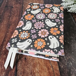 May include: A black book cover with a white and pink floral pattern and white ghost silhouettes. The cover has a white ribbon bookmark.