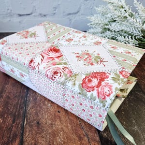 K&ouml;nnte beinhalten: Ein Buchumschlag aus floralem Stoff mit Patchwork-Design. Der Umschlag zeigt Rosen in Rosa und Rot sowie gr&uuml;n-wei&szlig;e Streifen. Ein gr&uuml;nes Leseb&auml;ndchen ist sichtbar.