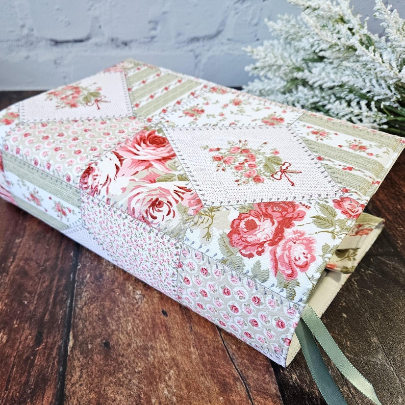 K&ouml;nnte beinhalten: Ein Buchumschlag aus floralem Stoff mit Patchwork-Design. Der Umschlag zeigt Rosen in Rosa und Rot sowie gr&uuml;n-wei&szlig;e Streifen. Ein gr&uuml;nes Leseb&auml;ndchen ist sichtbar.