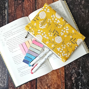 May include: A yellow fabric zippered pouch with a white floral pattern. The pouch is lying open on a book with colorful sticky notes and two multi-colored pens.