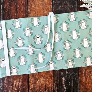 Peut inclure: Une housse de livre en tissu turquoise avec une fermeture en ruban blanc. Le tissu pr&eacute;sente un motif r&eacute;p&eacute;titif de fant&ocirc;mes blancs dans des couronnes florales.