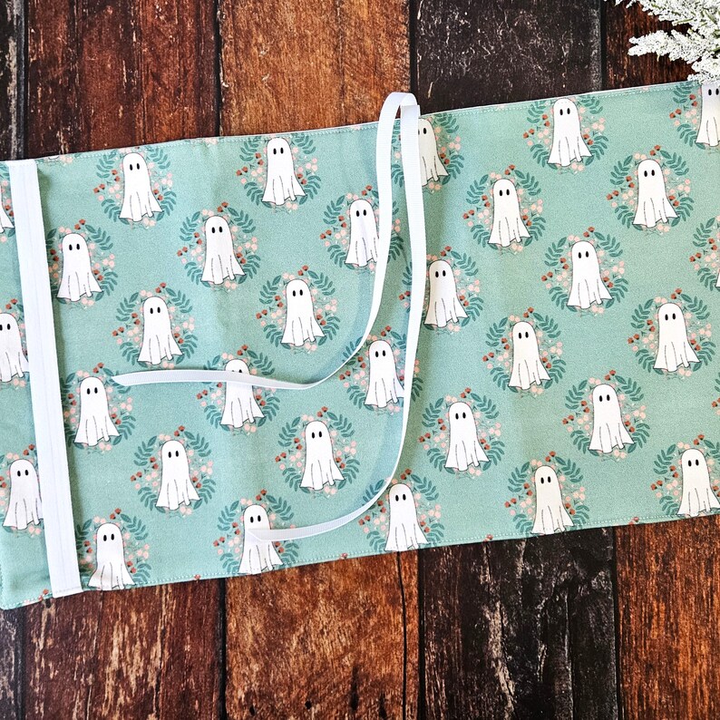 Peut inclure: Une housse de livre en tissu turquoise avec une fermeture en ruban blanc. Le tissu pr&eacute;sente un motif r&eacute;p&eacute;titif de fant&ocirc;mes blancs dans des couronnes florales.