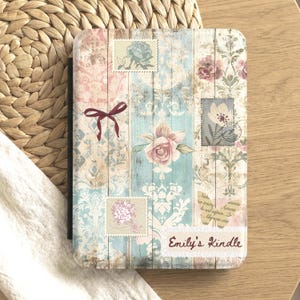 May include: Kindle cover with a floral and postage stamp design. The cover features a mix of pastel colors, including pink, blue, and cream, with a wooden plank background. A burgundy bow is on the upper left. The text "Emily's Kindle" is at the bottom.