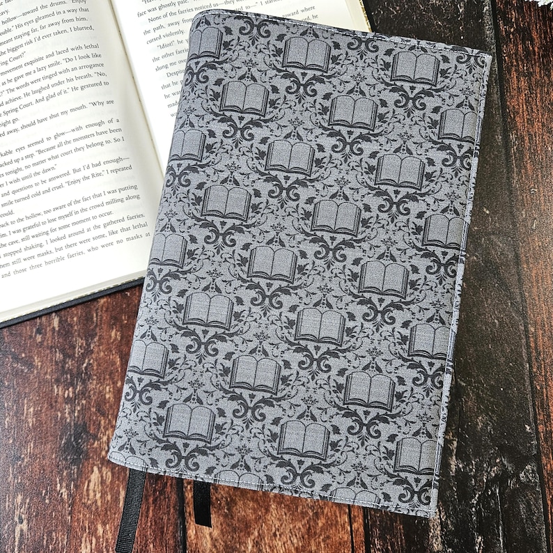 May include: A gray book cover with a repeating pattern of black open books and ornate designs. The cover has two black ribbon bookmarks.