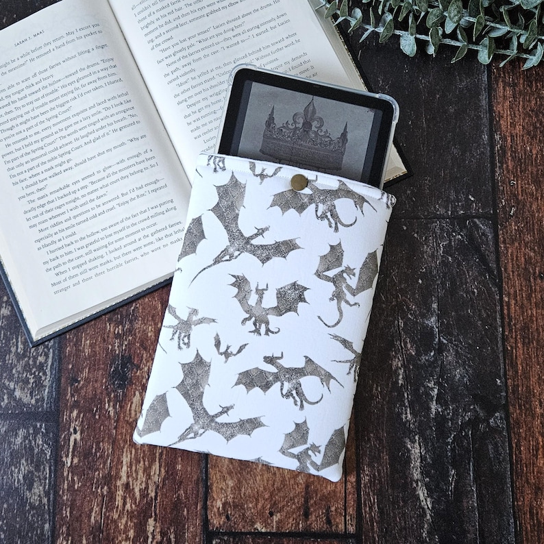 K&ouml;nnte beinhalten: Wei&szlig;e E-Reader-H&uuml;lle mit einem sich wiederholenden Muster aus grauen Drachen-Silhouetten. Die H&uuml;lle hat einen Knopfverschluss und ist mit einem E-Reader darin abgebildet.