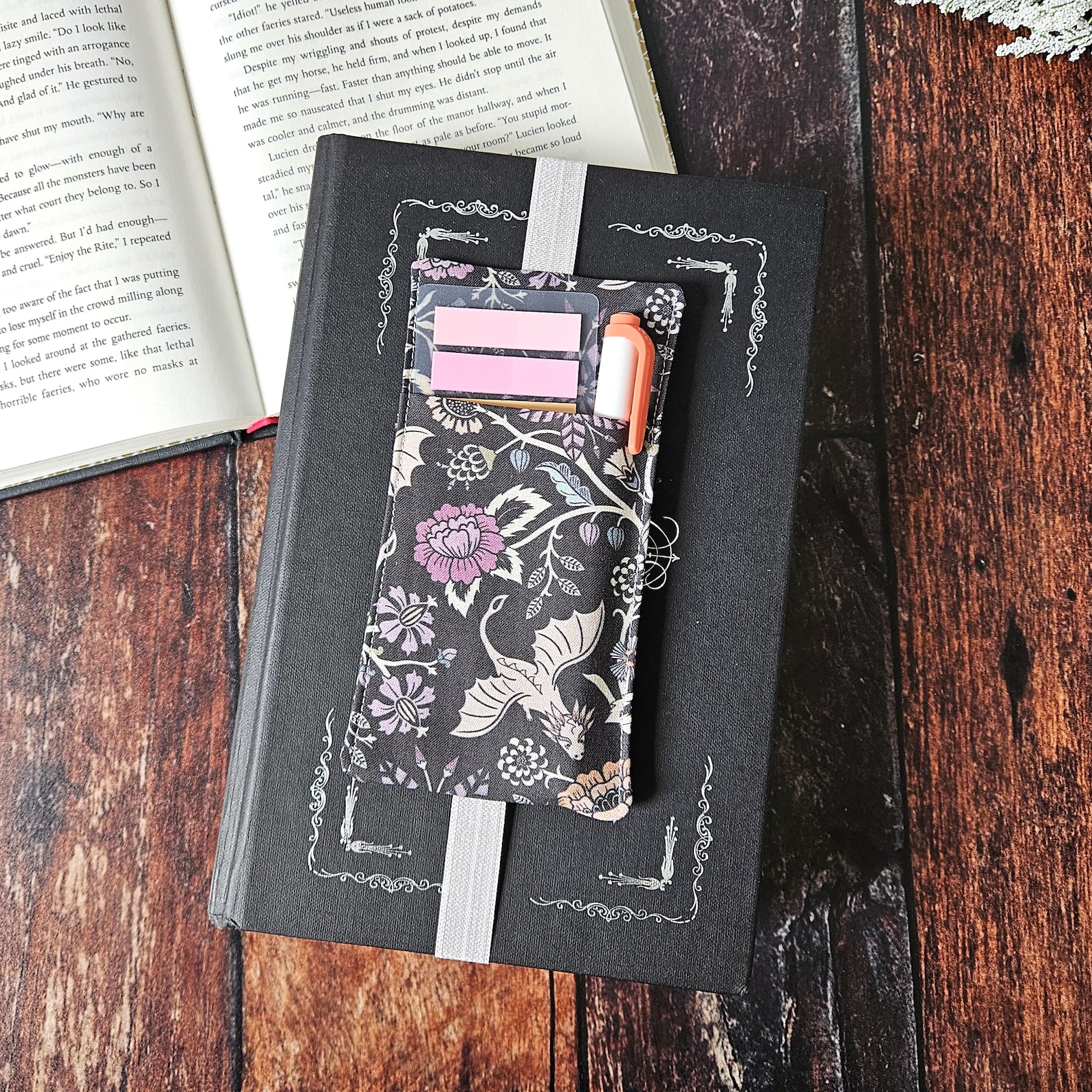A back hardcover book sitting on a wooden background, with a cloth pouch with dragons and flowers attached by an elastic band to the cover. Inside the pouch are sticky notes and a highlighter.