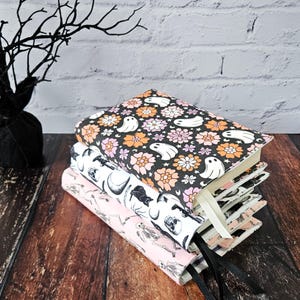 May include: A stack of three book covers with a Halloween theme. The top cover features a black background with white ghosts and pink and orange flowers. The middle cover features a black cat, a skull, and a crescent moon on a white background. The bottom cover features a pink background with a skeleton and a skull on a white background.