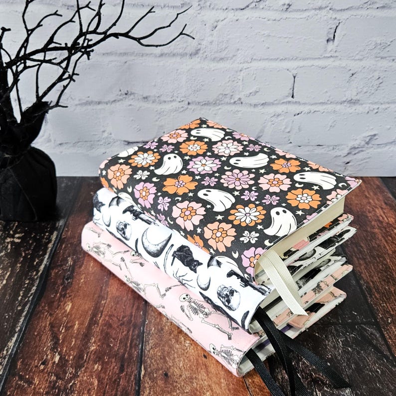 May include: A stack of three book covers with a Halloween theme. The top cover features a black background with white ghosts and pink and orange flowers. The middle cover features a black cat, a skull, and a crescent moon on a white background. The bottom cover features a pink background with a skeleton and a skull on a white background.