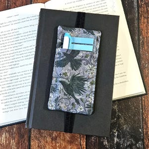 Puede incluir: Una funda de libro de tela con un diseño de cuervo, que contiene notas adhesivas azules y un bolígrafo. La funda se coloca encima de un libro negro cerrado con una banda elástica negra. También se ve un libro abierto con texto.