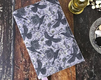 May include: A rectangular fabric book cover featuring a repeating pattern of black crows in flight, interspersed with white flowers and dark swirling vine motifs on a light grey background, with two light purple ribbon bookmarks attached.