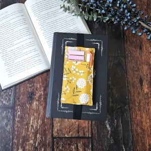 Op de afbeelding: Een gele stoffen boekomslag met een wit bloemenpatroon. De omslag heeft een vak voor een pen en plakbriefjes. De omslag ligt op een zwart boek.