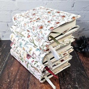 K&ouml;nnte beinhalten: Ein Stapel von vier Hardcover-B&uuml;chern mit floralen Stoffeinb&auml;nden. Jedes Buch hat ein anderes Blumenmuster in Rosa-, Rot- und Gr&uuml;nt&ouml;nen auf wei&szlig;em Hintergrund. Jedes Buch hat ein unterschiedlich farbiges Lesezeichenband.