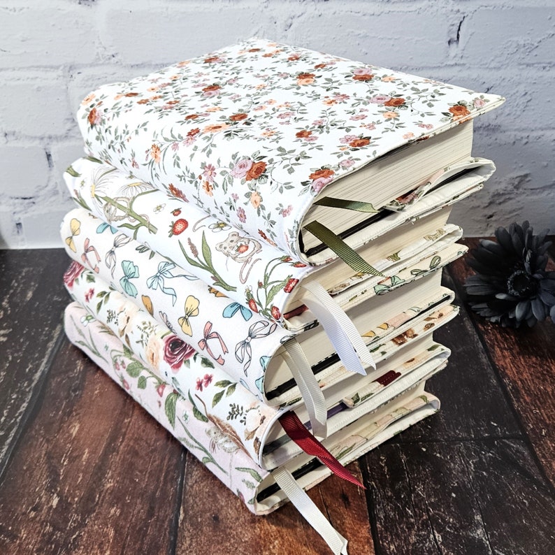 K&ouml;nnte beinhalten: Ein Stapel von vier Hardcover-B&uuml;chern mit floralen Stoffeinb&auml;nden. Jedes Buch hat ein anderes Blumenmuster in Rosa-, Rot- und Gr&uuml;nt&ouml;nen auf wei&szlig;em Hintergrund. Jedes Buch hat ein unterschiedlich farbiges Lesezeichenband.