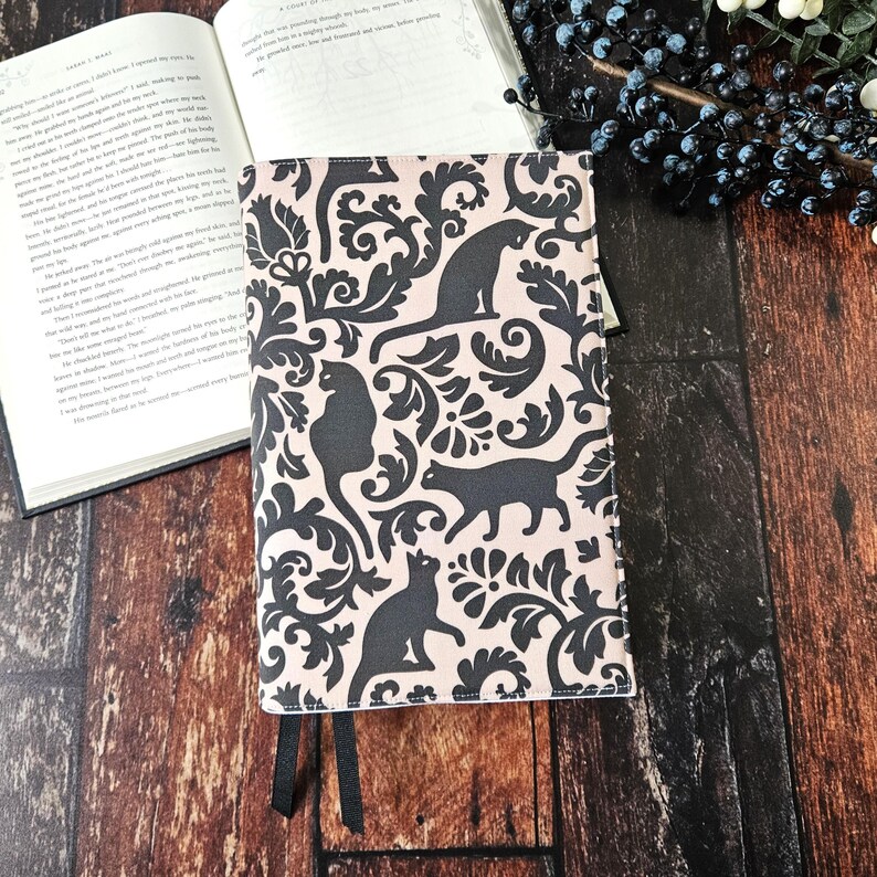 May include: A pink book cover with a black and white damask pattern featuring black cats. The cover has two black ribbon bookmarks.