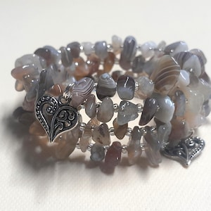 Può includere: Bracciale a più fili con pietre naturali burattate nei toni del grigio, marrone e crema. Il bracciale è impreziosito da due ciondoli a forma di cuore argentati. Le pietre sono infilate su un filo flessibile.