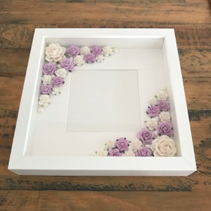 Puede incluir: Marco de fotos blanco con un paspart&uacute; blanco y un fondo blanco. El marco est&aacute; decorado con flores moradas y blancas.