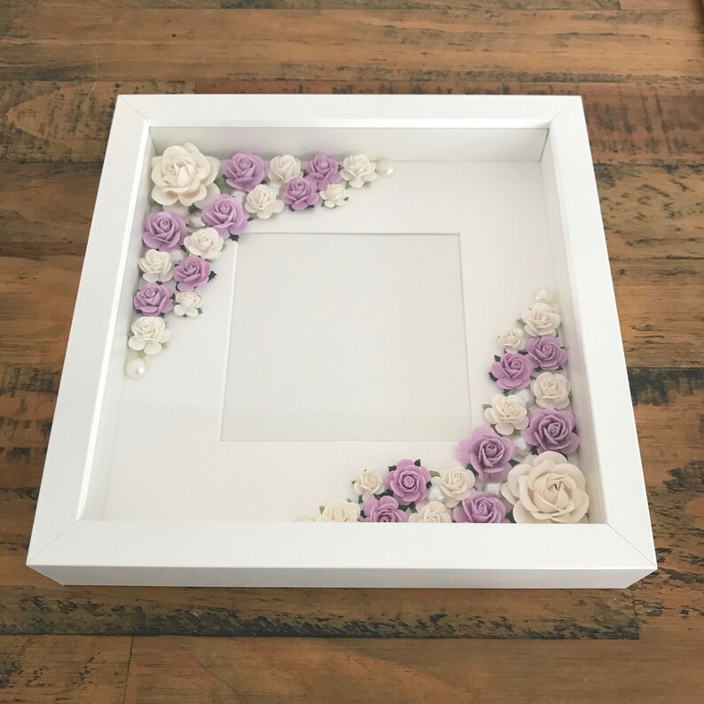 Puede incluir: Marco de fotos blanco con un paspart&uacute; blanco y un fondo blanco. El marco est&aacute; decorado con flores moradas y blancas.