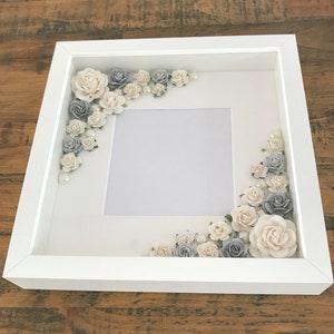 Puede incluir: Marco de fotos blanco con un paspart&uacute; blanco y un fondo blanco. El marco est&aacute; decorado con flores blancas y grises y perlas.