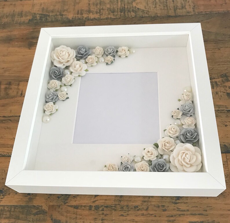 Puede incluir: Marco de fotos blanco con un paspart&uacute; blanco y un fondo blanco. El marco est&aacute; decorado con flores blancas y grises y perlas.