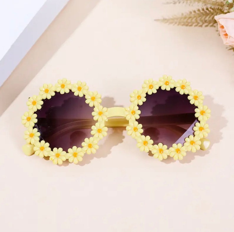 May include: Yellow sunglasses with a daisy flower design around the lenses. The sunglasses have a dark lens.