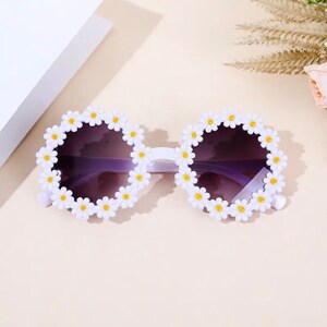 May include: White plastic sunglasses with a daisy design. The lenses are purple.