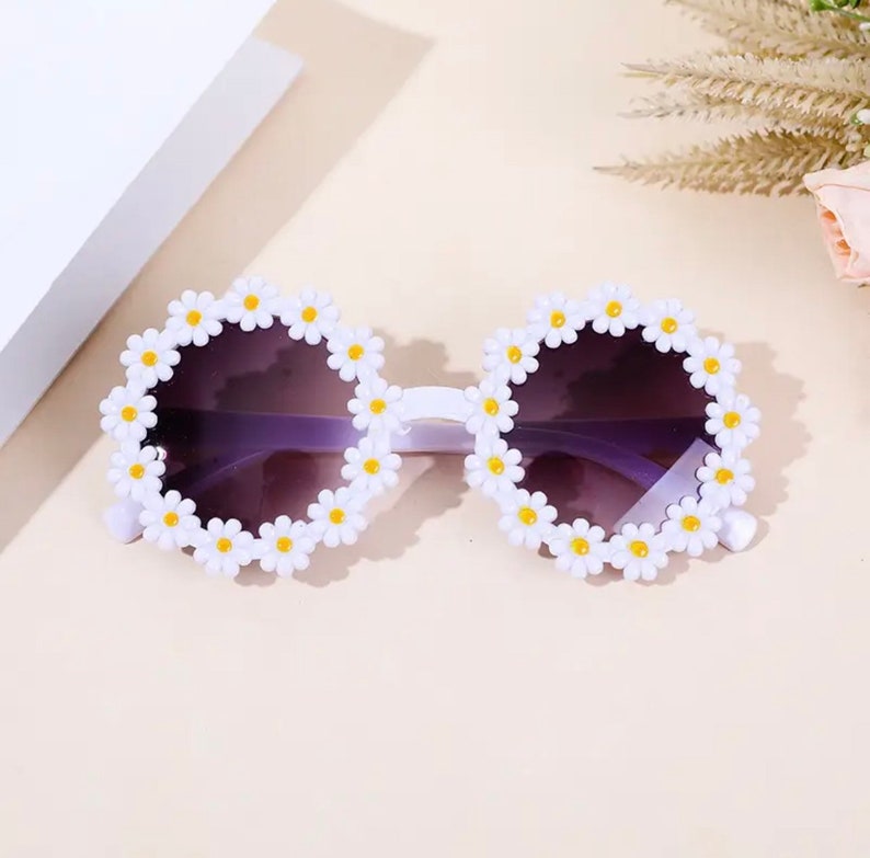 May include: White plastic sunglasses with a daisy design. The lenses are purple.