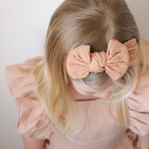 May include: A light brown headband with a large bow tied in the center. The headband is made of a soft, textured fabric.